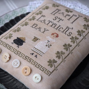 Puede incluir: Una almohada beige con un diseño de punto de cruz que representa a una niña sosteniendo una olla de oro y el texto "Feliz Día de San Patricio". La almohada está decorada con botones blancos.