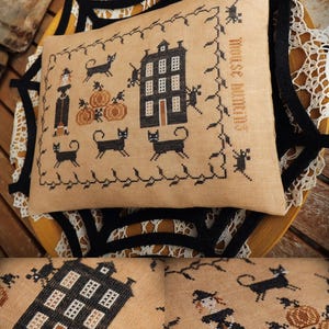 Op de afbeelding: Een beige kruissteek sampler met een Halloween thema. Het ontwerp toont een zwart huis, zwarte katten, pompoenen en een heks. De woorden "MOUSE HOUSE" zijn in oranje gestikt. De sampler is omlijst met een zwarte rankenrand.
