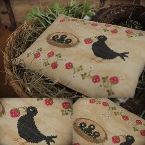 Puede incluir: Un cojín decorativo cuadrado beige con un diseño de punto de cruz que presenta un pájaro negro, un nido con tres polluelos y fresas rojas con flores blancas. El cojín está en una cesta tejida llena de heno.