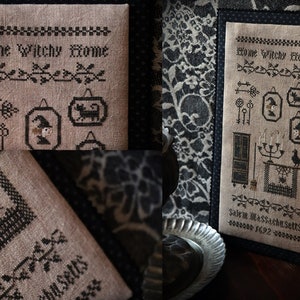 Könnte beinhalten: Ein Kreuzstich-Sampler mit dem Text "Home Witchy Home" und verschiedenen Hexen-Motiven, darunter ein Besen, ein Kessel, eine schwarze Katze und ein Hexenhut. Der Sampler ist in einem schwarzen Rahmen gerahmt und vor einem schwarz-weißen floralen Hintergrund platziert. Der Text "Salem, Massachusetts 1692" ist ebenfalls im Sampler enthalten.