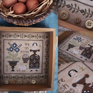May include: A framed cross-stitch sampler with the words "fresh eggs" and a woman in a brown dress. A basket of brown eggs sits above the sampler. Several buttons are placed below the sampler.