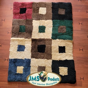 May include: A colorful sheepskin rug with a geometric pattern of squares in shades of red, brown, green, black, cream, and blue. The rug has a "JMS Products Just Merino Sheepskin" logo in the bottom right corner.