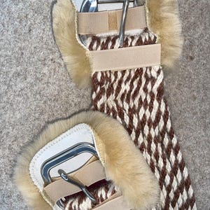 May include: A close-up of a horse cinch. The cinch features a woven brown and white pattern with tan fur padding around the edges. It has a silver buckle and tan straps.