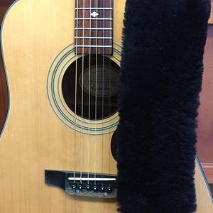 May include: Acoustic guitar with a natural wood finish, featuring a black bridge and strings. A black, fluffy cover is placed over the neck. The guitar has a sound hole with concentric rings and a brand label.