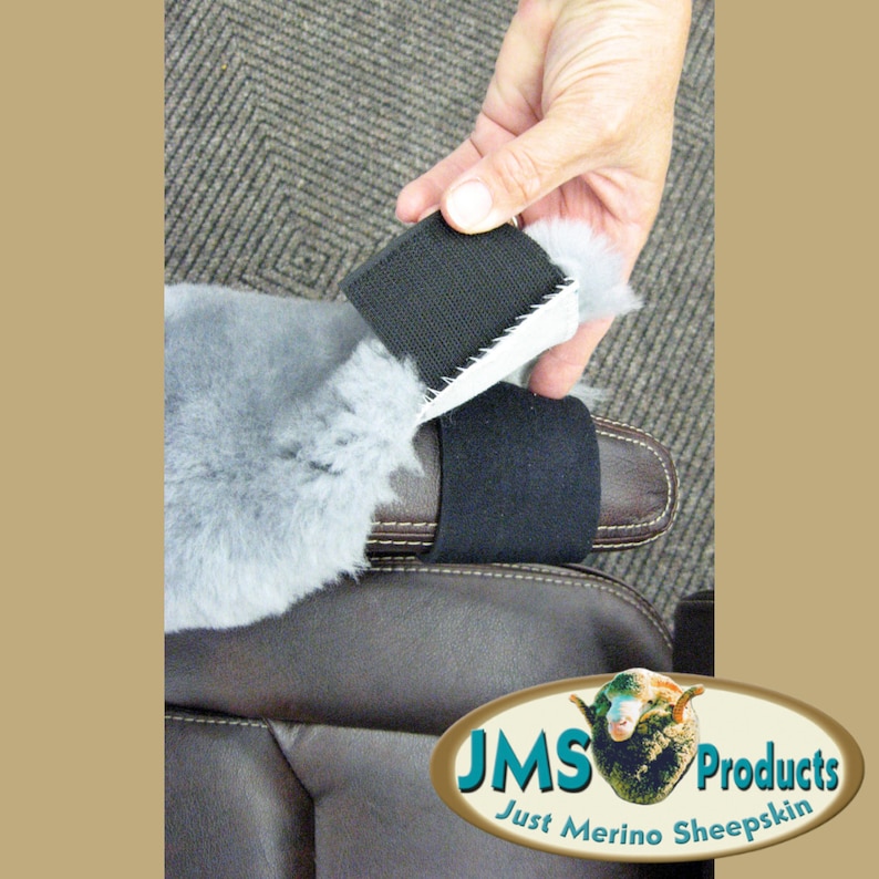 May include: A close-up of a gray sheepskin car seat cover with a black strap. The strap is attached to the cover with a hook and loop fastener. The cover is being held up by a hand.