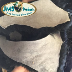 May include: Close-up of a black sheepskin with the white leather side turned up, showing the fleece. The sheepskin is labeled "JMS Products Just Merino Sheepskin".