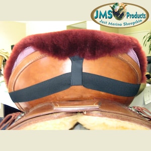 May include: A burgundy sheepskin saddle pad with a black elastic strap. The pad is on a brown leather saddle.