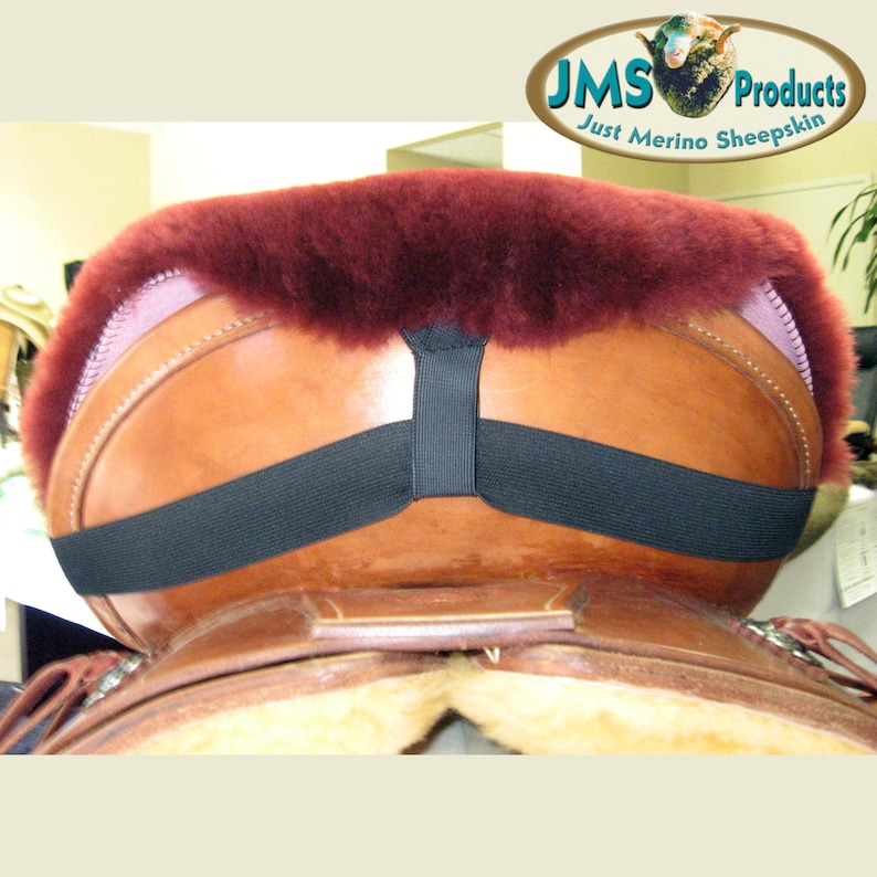 May include: A burgundy sheepskin saddle pad with a black elastic strap. The pad is on a brown leather saddle.