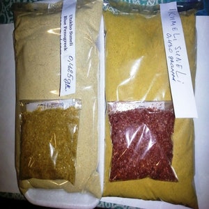 Może przedstawiać: Dwie przezroczyste plastikowe torby wypełnione żółtymi i beżowymi proszkami, oznaczone jako "Uskho Suneli Blue Fenugreek" i "Ikumeli Suneli". Każda torba zawiera mniejszy pakiet innej przyprawy. Torby są zamknięte i gotowe do użycia.