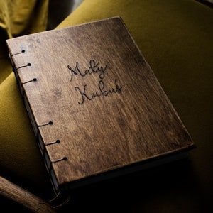 Op de afbeelding: Een houten dagboek met de naam "Maly Kubus" in cursiefschrift. Het dagboek is gebonden met zichtbare stiksels en ligt op een groene gestoffeerde stoel. Het hout heeft een warme, natuurlijke nerf.