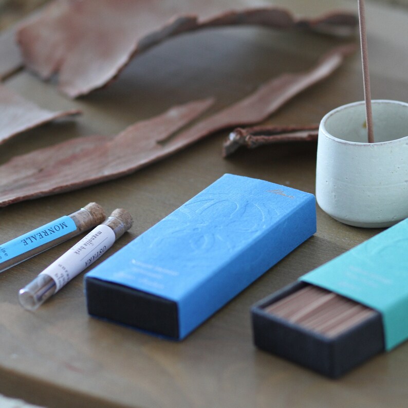 May include: Two boxes of incense sticks, one blue and one teal, with a label that reads "COSMIC" and "MONREALE" on the blue box. The boxes are on a wooden surface with a piece of bark and a small white ceramic cup.