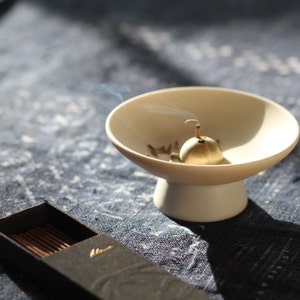 May include: A white ceramic incense burner with a gold metal incense holder. The incense holder is shaped like a small bell and has a small hole in the top. Smoke is rising from the incense holder.