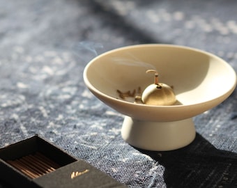 May include: A white ceramic incense burner with a gold metal incense holder. The incense holder is shaped like a small bell and has a small hole in the top. Smoke is rising from the incense holder.