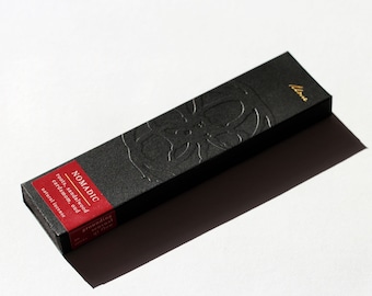 May include: Black box with gold lettering that reads "NOMADIC" and "grounding essential oil" with a description of the scent: "roots, sandalwood, cardamom, oud, natural incense". The box is embossed with a floral design and has the word "blume" in gold lettering on the top right corner.
