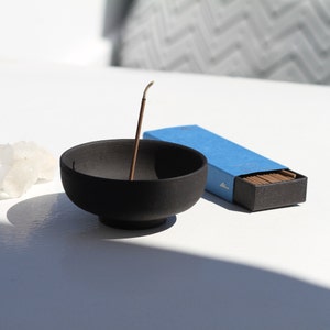 May include: A black ceramic incense holder with a single incense stick burning. The incense stick is in a blue box with the word "Hina" printed on it.