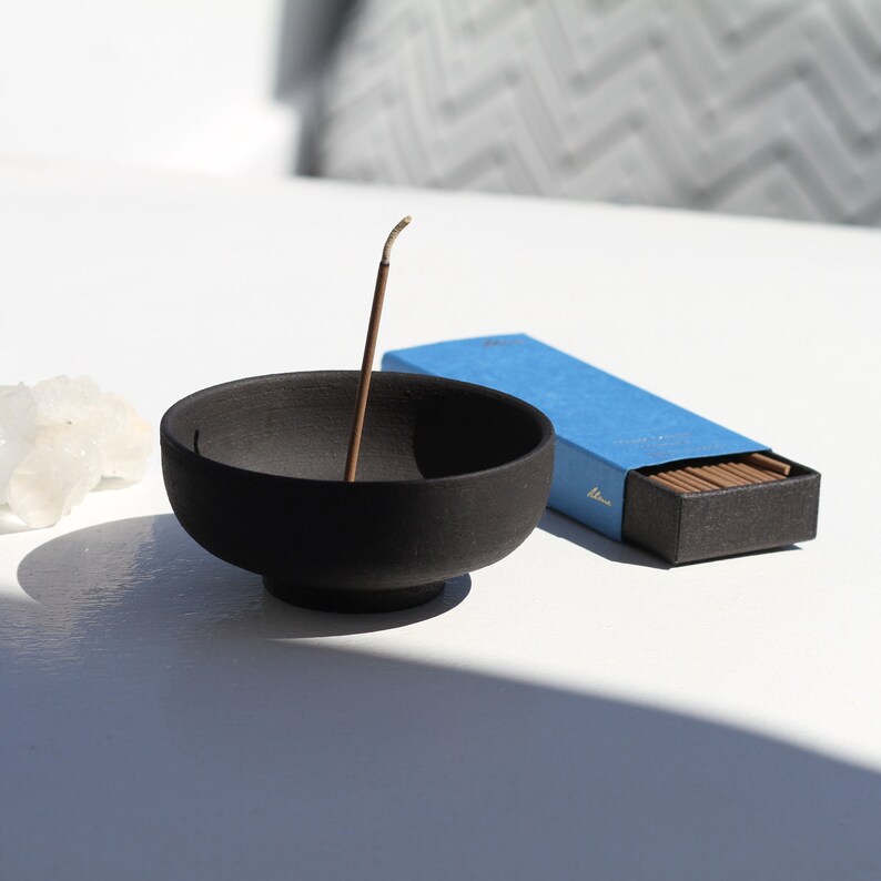 May include: A black ceramic incense holder with a single incense stick burning. The incense stick is in a blue box with the word "Hina" printed on it.