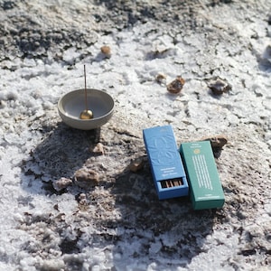May include: Two boxes of incense sticks on a white, rocky surface. The boxes are blue and green with gold lettering. The text on the blue box reads "Sacred Smoke Almond Blossom Agarbathi". The text on the green box reads "Wild Herbs & Spices Agarbathi". A small gray ceramic incense holder with a gold ball on top is in the background.