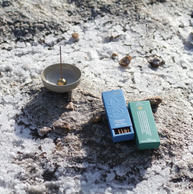 May include: Two boxes of incense sticks on a white, rocky surface. The boxes are blue and green with gold lettering. The text on the blue box reads "Sacred Smoke Almond Blossom Agarbathi". The text on the green box reads "Wild Herbs & Spices Agarbathi". A small gray ceramic incense holder with a gold ball on top is in the background.