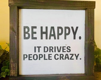 May include: A wooden sign with a white background and black text that reads "BE HAPPY. IT DRIVES PEOPLE CRAZY."