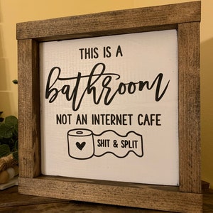 May include: A white wooden sign with a brown frame reads "This is a bathroom, not an internet cafe. Shit & Split." A black and white illustration of a toilet paper roll with a heart on it is below the text.