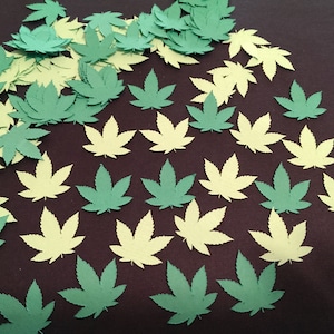 Peut inclure: Une collection de découpes de feuilles de cannabis en papier vert et jaune. Les feuilles sont disposées en motif sur une surface brun foncé.