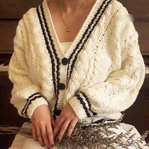 Könnte beinhalten: Ein weißer Strickcardigan mit schwarzem Besatz und Knopfleiste. Der Cardigan hat ein Zopfmuster und einen V-Ausschnitt.