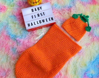 May include: An orange crochet baby outfit, including a hat and a cocoon, with a jack-o'-lantern and a sign that reads "BABY FIRST HALLOWEEN." The items are displayed on a colorful, fluffy surface.
