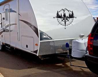 Peut inclure: Une caravane blanche avec un autocollant noir représentant une boussole et des montagnes sur le côté. La boussole indique le nord, le sud, l'est et l'ouest. L'autocollant représente une chaîne de montagnes avec des arbres.