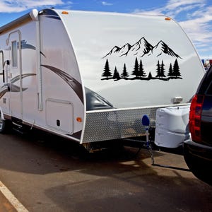 May include: A white camper with a black mountain and pine tree decal on the side. The camper is parked on a paved surface.