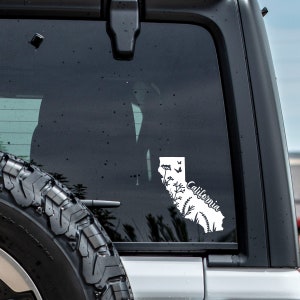May include: White vinyl decal of the state of California with a floral design and the word "California" in cursive. The decal is on the back window of a vehicle.