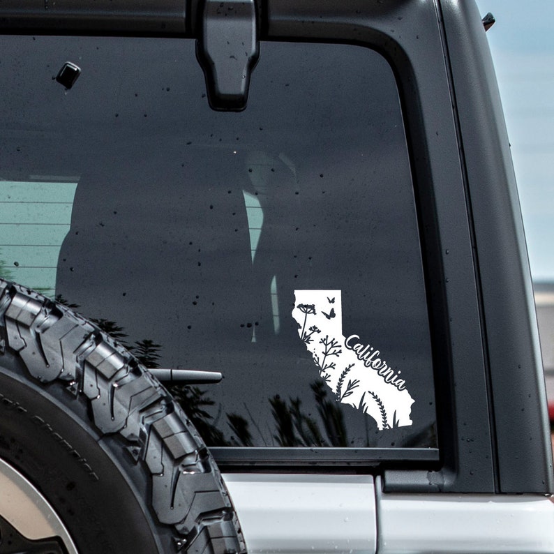 May include: White vinyl decal of the state of California with a floral design and the word "California" in cursive. The decal is on the back window of a vehicle.