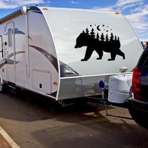 May include: A white camper trailer with a black bear silhouette decal on the side. The decal features a forest scene with trees and a crescent moon. The camper is parked on a paved surface.