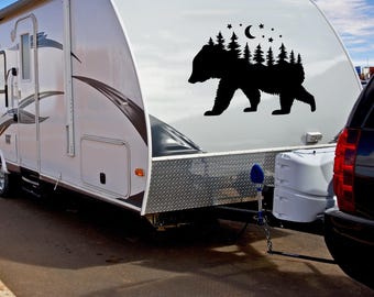 May include: A white camper trailer with a black bear silhouette decal on the side. The decal features a forest scene with trees and a crescent moon. The camper is parked on a paved surface.