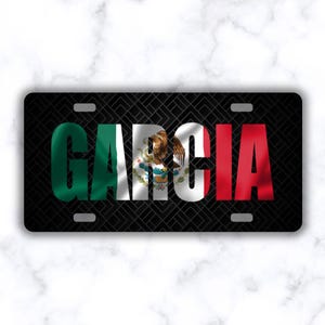 May include: Black license plate with the name "GARCIA" in white letters. The letters are filled with the colors of the Mexican flag: green, white, and red. The Mexican coat of arms is in the letter "C".