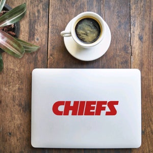 Pode incluir: Um laptop branco com um adesivo de logotipo "CHIEFS" vermelho na tampa. O laptop est&aacute; em uma mesa de madeira com uma x&iacute;cara de caf&eacute; e uma planta em vaso.
