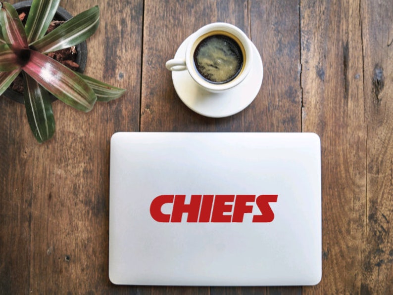 Pode incluir: Um laptop branco com um adesivo de logotipo "CHIEFS" vermelho na tampa. O laptop est&aacute; em uma mesa de madeira com uma x&iacute;cara de caf&eacute; e uma planta em vaso.