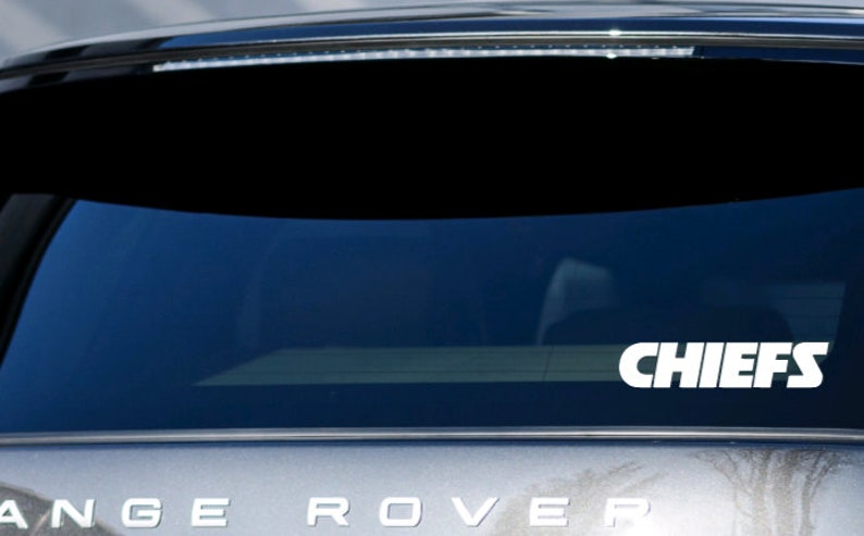 Pode incluir: Um adesivo de vinil branco com a palavra "CHIEFS" em letras brancas e negrito sobre um fundo preto. O adesivo est&aacute; na janela traseira de um carro prateado.