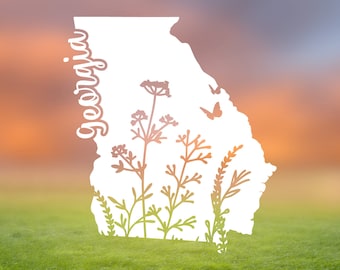 Peut inclure: Une silhouette blanche de l'état de Géorgie avec le mot « Géorgie » écrit verticalement sur le côté gauche. La silhouette est remplie d'une silhouette de différents types de plantes et de fleurs. La silhouette est placée sur un fond flou d'un ciel de coucher de soleil et d'un champ d'herbe verte.