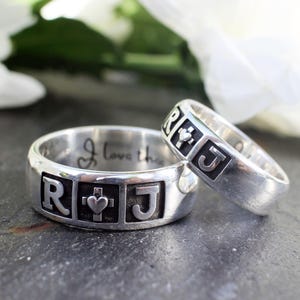 May include: Two silver rings with engraved letters and a cross with a heart. The ring on the left says "R + J" and the ring on the right says "R + J".