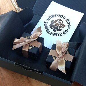 May include: Two small black gift boxes with gold ribbon bows. The boxes are inside a larger black box. The smaller boxes have a gold logo that says "Burning Rose Jewellery Co."