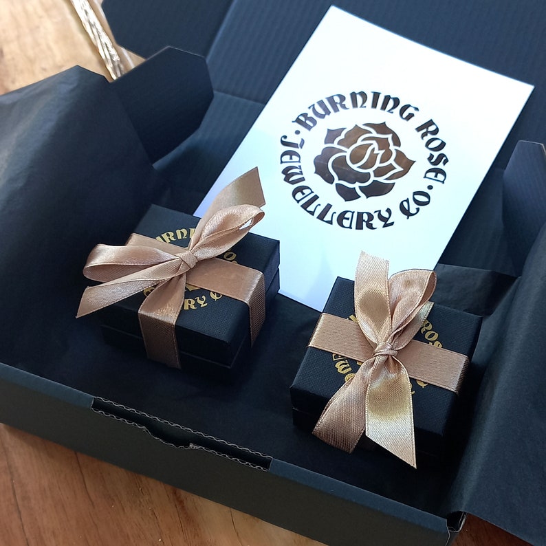 May include: Two small black gift boxes with gold ribbon bows. The boxes are inside a larger black box. The smaller boxes have a gold logo that says "Burning Rose Jewellery Co."