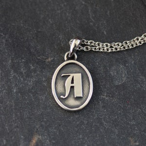 May include: Silver pendant necklace with an oval charm featuring a stylized letter A.