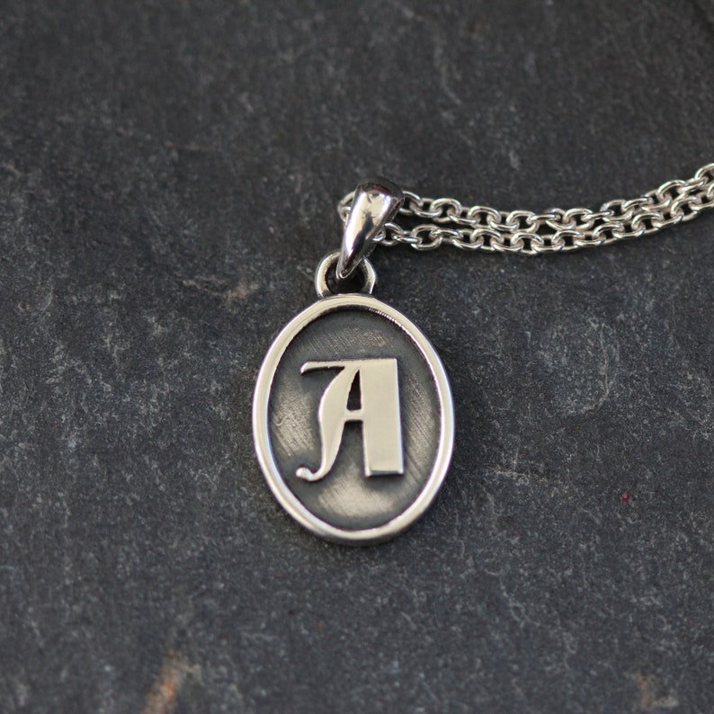 May include: Silver pendant necklace with an oval charm featuring a stylized letter A.