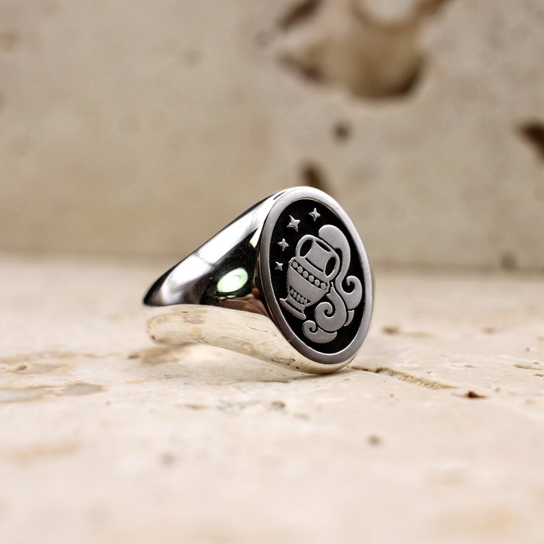 A silver, minimalist, oval signet ring with a tilted roman style vase, overflows with pouring water. In the black background are 4 little stars in the sky.