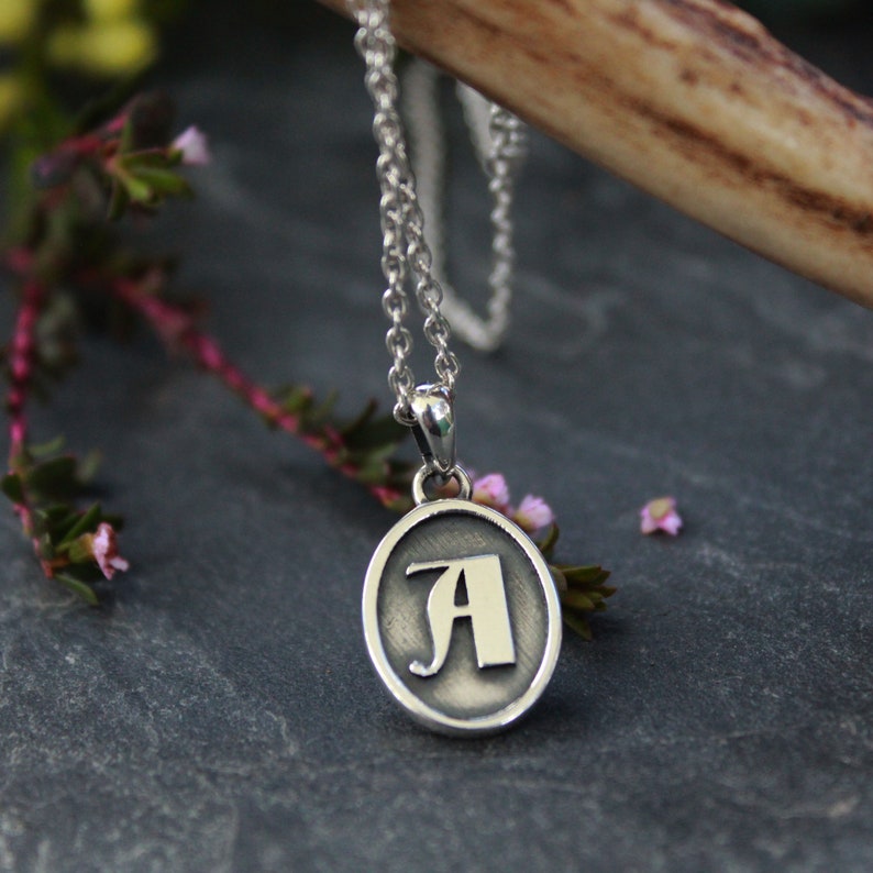 May include: A silver chain necklace with a silver oval pendant featuring the letter 'A' in a stylized font.