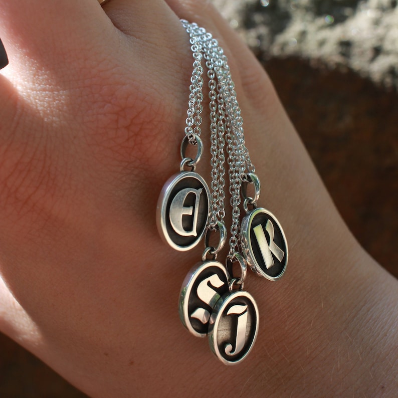 May include: Five silver necklaces with round pendants featuring the letters E, S, J, and K. The necklaces are layered and hanging from a person's hand.
