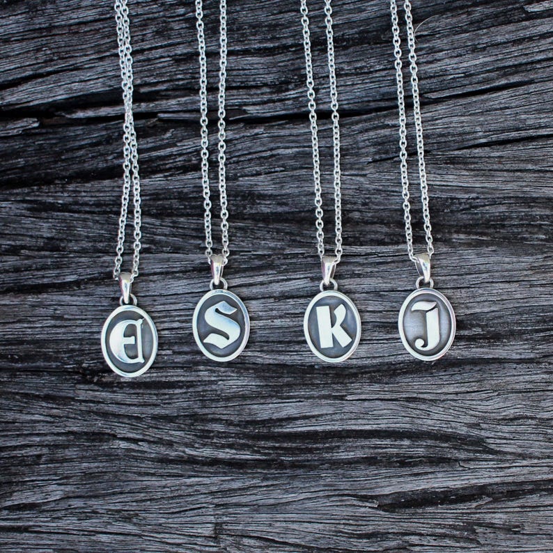 May include: Five silver necklaces with oval pendants featuring the letters E, S, K, and J in a gothic font.