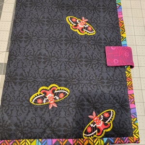 May include: A black fabric folder with a colorful border and three pink and yellow moths. The folder has a pink fabric flap with a black and white pattern.