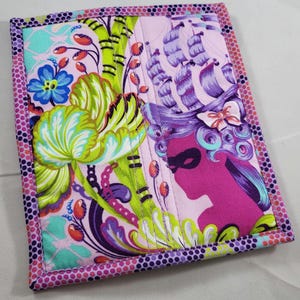 May include: A rectangular quilted fabric piece featuring a vibrant print of a pink silhouette wearing a ship-shaped hat, alongside large green and yellow floral motifs on a light pink background, finished with a purple and orange polka dot border.