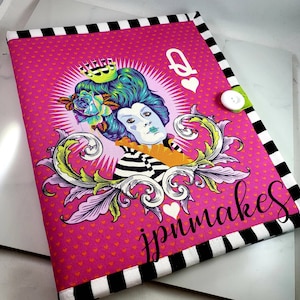 May include: A pink and black fabric notebook cover with a white button. The cover features a whimsical illustration of a queen with a crown, a floral design, and the text "jpmnakes".